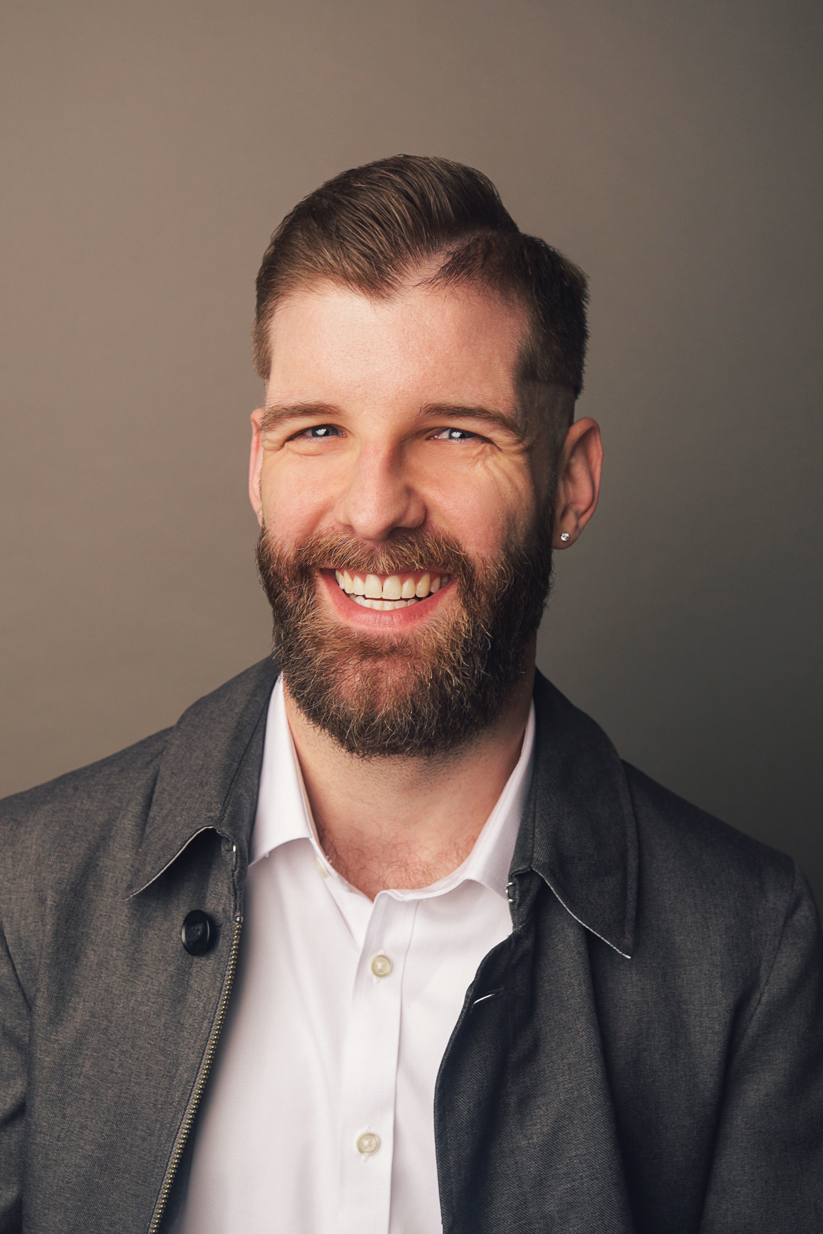 A portrait photograph of Michael Walker. He wears a gray coat and white dress shirt on a beige gradient background. His hair is clean and neat and he smiles warmly. His blue eyes face the viewer with a twinkle.